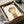 Minimalist illustration of a young child with short brown hair and a solemn expression, wearing an orange sleeveless top and holding a small green sprout. The artwork features soft, muted tones on cream paper, displayed in a black frame being unpacked from a cardboard shipping box with protective tissue paper. The gentle, cartoon-like style emphasizes the child's downward gaze and peaceful demeanor.