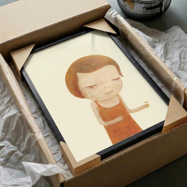 Minimalist illustration of a young child with short brown hair and a solemn expression, wearing an orange sleeveless top and holding a small green sprout. The artwork features soft, muted tones on cream paper, displayed in a black frame being unpacked from a cardboard shipping box with protective tissue paper. The gentle, cartoon-like style emphasizes the child's downward gaze and peaceful demeanor.
