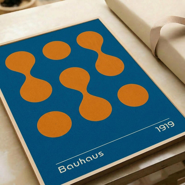May include: Blue poster with abstract orange geometric shapes resembling connected circles and organic forms, displayed on white paper with ribbon nearby. Clean minimalist design with "Bauhaus" and "1919" text at bottom in white lettering against the vibrant blue background.