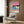 May include: Framed poster showing tropical beach scene with pink sunset sky, palm trees, blue ocean waves, and two small figures on sandy shore. Displayed on wood-paneled wall above cream upholstered armchair with geometric patterned pillow. Modern brass floor lamp and dark round side table with open book complete the minimalist living room setting with light wood flooring and white curtains.
