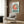 May include: Studio Ghibli Ponyo poster mounted on wood-paneled wall above beige upholstered armchair with geometric pillow. Poster features red-haired Ponyo character in circular blue frame with "PONYO" text below. Modern living room setting with brass floor lamp, side table with open book, and natural lighting from window with curtains.