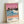 May include: Framed art print leaning against white wall showing tropical beach scene with pink sunset sky, palm trees on hillside, blue ocean waves with white foam, coral-pink sand, and two small silhouetted figures. Features artist name "Hiroshi Nagai" and "Tokyo 1973" text at bottom with colorful dots. Light wood frame, soft natural lighting creates shadows on wall.