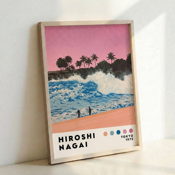 May include: Framed art print leaning against white wall showing tropical beach scene with pink sunset sky, palm trees on hillside, blue ocean waves with white foam, coral-pink sand, and two small silhouetted figures. Features artist name "Hiroshi Nagai" and "Tokyo 1973" text at bottom with colorful dots. Light wood frame, soft natural lighting creates shadows on wall.