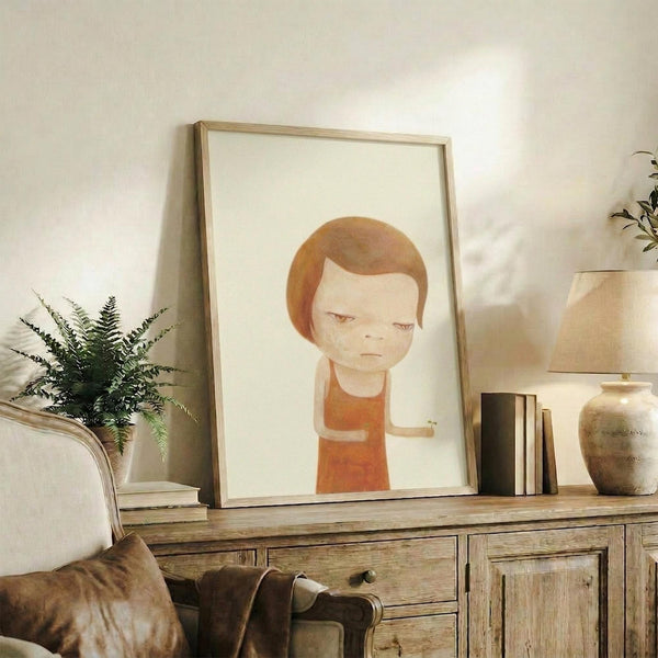 May include: Framed art print featuring a stylized illustration of a young child with orange-brown hair and closed eyes, wearing an orange-red shirt, displayed on a rustic wooden sideboard alongside decorative pottery, books, and a fern plant in a cozy living room setting with neutral tones and soft lighting.