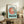 May include: Framed Studio Ghibli Ponyo poster displayed on rustic wooden sideboard in cozy living room setting. The artwork features the red-haired character Ponyo in a circular design with turquoise background and brown lettering. Natural wood frame complements surrounding decor including fern plant, ceramic vase, stacked books, and neutral-toned furniture with throw pillows.