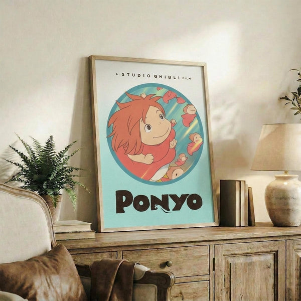 May include: Framed Studio Ghibli Ponyo poster displayed on rustic wooden sideboard in cozy living room setting. The artwork features the red-haired character Ponyo in a circular design with turquoise background and brown lettering. Natural wood frame complements surrounding decor including fern plant, ceramic vase, stacked books, and neutral-toned furniture with throw pillows.