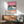 May include: Framed poster with tropical beach scene in pink sunset sky, blue ocean waves, palm tree silhouettes, and small figures on coral sand. Displayed above mid-century modern wooden credenza with white ceramic lamp, next to sage green upholstered armchair with wood frame. Bright natural light streams across white wall creating geometric shadows.