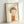 May include: Framed art print leaning against white wall showing minimalist illustration of a child-like figure with brown bob haircut and orange-red clothing on cream background. Light wood frame casts soft shadows on white surface below.