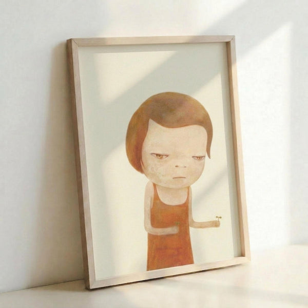 May include: Framed art print leaning against white wall showing minimalist illustration of a child-like figure with brown bob haircut and orange-red clothing on cream background. Light wood frame casts soft shadows on white surface below.