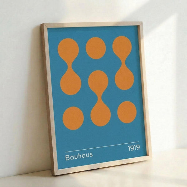 A geometric poster with orange circles and connecting shapes arranged in a 3x3 grid pattern on a bright blue background. The text "Bauhaus 1919" appears in white at the bottom. The artwork is displayed in a thin light wood frame and leaning against a white wall, casting a soft shadow.