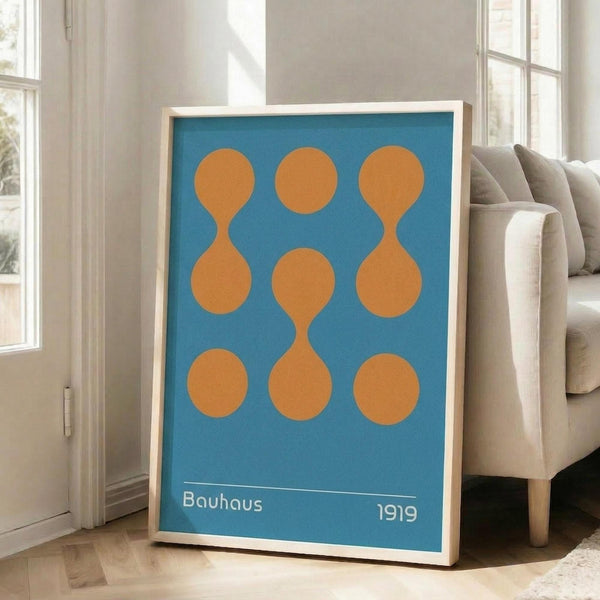 May include: A framed geometric poster with blue background and nine orange circular shapes arranged in a 3x3 grid, some connected to form hourglass or figure-eight patterns. White text reads "Bauhaus" and "1919" at bottom. Light wood frame leans against white wall next to beige upholstered sofa in bright, minimalist living room with large windows.