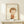 May include: Framed art print featuring a stylized illustration of a young child with brown bowl-cut hair wearing an orange shirt, rendered in soft watercolor-like tones against a cream background. The wooden frame sits on a light wood surface alongside a small white gift box with evergreen sprig, creating a minimalist holiday display against white walls.