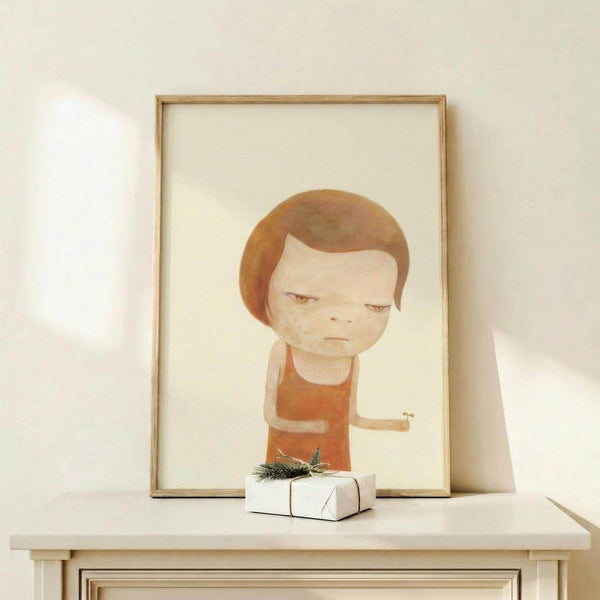 May include: Framed art print featuring a stylized illustration of a young child with brown bowl-cut hair wearing an orange shirt, rendered in soft watercolor-like tones against a cream background. The wooden frame sits on a light wood surface alongside a small white gift box with evergreen sprig, creating a minimalist holiday display against white walls.