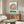 May include: Studio Ghibli Ponyo movie poster print displayed above a mid-century modern wooden credenza in a minimalist living room. The poster features the red-haired character Ponyo in a circular frame against a turquoise background with black text below. Room has cream walls, white modern chair, brass floor lamp, oval glass coffee table, and white table lamp on the credenza.