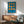 May include: Framed Bauhaus-style geometric poster with blue background and orange circular shapes arranged in a 3x3 grid, displayed above a wooden mid-century modern credenza with table lamp, next to a teal upholstered armchair with wooden frame in a sunlit room with white walls and hardwood floors.