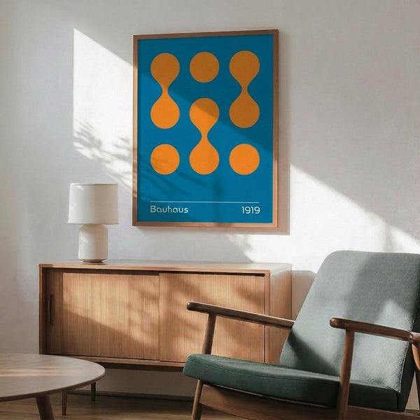 May include: Framed Bauhaus-style geometric poster with blue background and orange circular shapes arranged in a 3x3 grid, displayed above a wooden mid-century modern credenza with table lamp, next to a teal upholstered armchair with wooden frame in a sunlit room with white walls and hardwood floors.