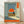 May include: Framed poster print leaning against white wall on wooden floor. Artwork shows person sitting in field viewing large orange sun and clouds through doorway or window frame. Japanese text in orange banner on right side. Vintage-inspired illustration with muted blue-green and orange color palette. Bottom has orange text block. Natural lighting creates soft shadows on wall.