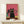 May include: Framed poster print with pink background showing group of cats wearing sunglasses, with prominent black cat in center wearing yellow collar. Various colored cats including teal, pink, and black positioned behind main figure. Thin gold frame displayed on light wood surface next to small wrapped gift with twine and greenery sprig against cream wall.