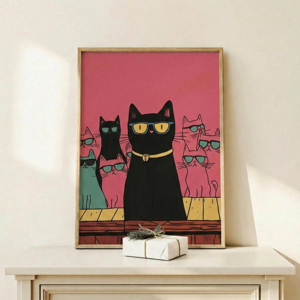 May include: Framed poster print with pink background showing group of cats wearing sunglasses, with prominent black cat in center wearing yellow collar. Various colored cats including teal, pink, and black positioned behind main figure. Thin gold frame displayed on light wood surface next to small wrapped gift with twine and greenery sprig against cream wall.