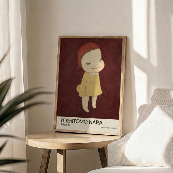 May include: Framed poster featuring a stylized illustration of a child-like figure with red hair wearing a yellow dress against a deep burgundy background, displayed on a round wooden side table next to a beige upholstered chair with a white pillow, green plant leaves visible in corner, soft natural lighting creating shadows on white wall