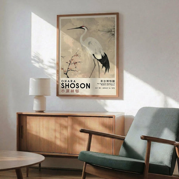 May include: Vintage Japanese crane art poster in wooden frame hanging above mid-century modern furniture. The print features a white heron with black wing tips and red crown detail, surrounded by delicate botanical elements on aged beige background. Museum exhibition text visible below the artwork. Displayed in minimalist living room with sage green armchair, wooden sideboard, ceramic table lamp, and natural sunlight casting shadows across white walls.