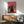 May include: Framed poster print featuring stylized cats wearing colorful sunglasses against a pink gradient background. The main black cat sits prominently in center with yellow sunglasses and collar, surrounded by smaller pink and teal cats also wearing glasses. Print displayed above a mid-century modern wooden sideboard with white ceramic lamp, next to a sage green upholstered armchair with wooden arms in a bright, minimalist living room setting.