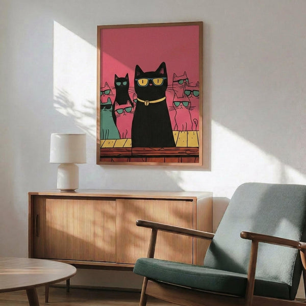 May include: Framed poster print featuring stylized cats wearing colorful sunglasses against a pink gradient background. The main black cat sits prominently in center with yellow sunglasses and collar, surrounded by smaller pink and teal cats also wearing glasses. Print displayed above a mid-century modern wooden sideboard with white ceramic lamp, next to a sage green upholstered armchair with wooden arms in a bright, minimalist living room setting.