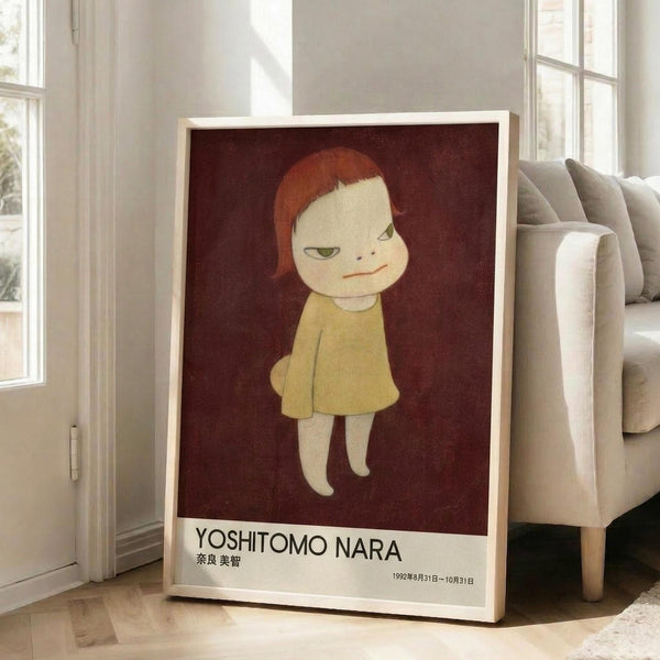 May include: Framed poster featuring a stylized illustration of a pale child-like figure with reddish-brown hair wearing a yellow dress against a deep burgundy background. The print has white text reading "YOSHITOMO NARA" and Japanese characters below, with exhibition dates. The wooden-framed artwork leans against a white wall next to a beige upholstered sofa in a bright, minimalist living room with large windows.