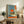 May include: Framed poster with Japanese aesthetic showing figure sitting before large orange sun over water, viewed through doorway. Blue and orange color palette with Japanese text. Displayed on rustic wooden console alongside fern plant, books, and ceramic vase in neutral-toned living space with beige chair visible.