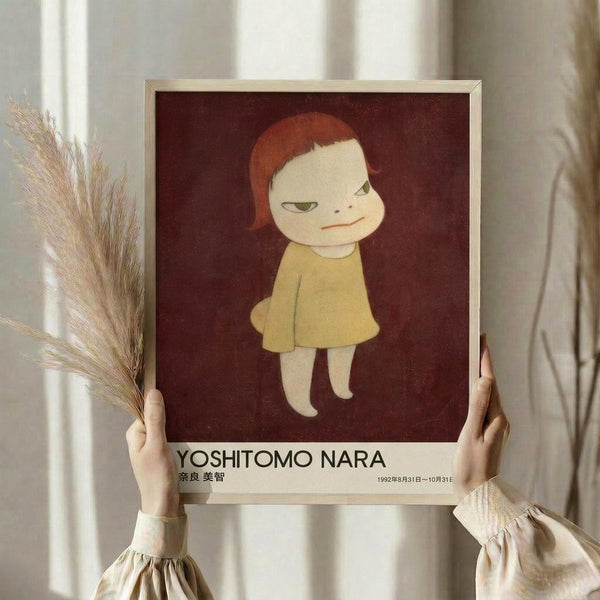 May include: Person holding framed art print featuring stylized illustration of child-like figure with red hair and yellow dress against burgundy background. White matted frame with "YOSHITOMO NARA" text below artwork. Person wears cream ruffled sleeves, holds dried pampas grass stems. Soft natural lighting creates shadows on neutral wall background.