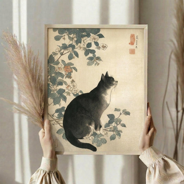 May include: Person in cream ruffled sleeves holding framed vintage Japanese artwork showing black cat sitting beneath flowering rose branches with green leaves. Person also holds dried wheat stems. Artwork has beige background with red seal stamps in traditional ukiyo-e style. Light wooden frame against soft neutral curtained background.
