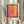 May include: Person holding framed art print featuring abstract color field painting with vibrant pink center, yellow right section, and teal-blue left border. Light wood frame with white mat displaying artist name and retrospective dates below artwork. Neutral beige curtain background with dried pampas grass visible on left side.