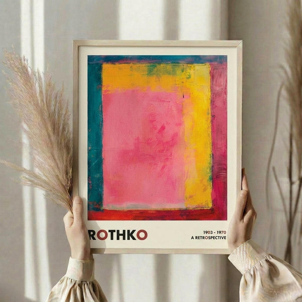 May include: Person holding framed art print featuring abstract color field painting with vibrant pink center, yellow right section, and teal-blue left border. Light wood frame with white mat displaying artist name and retrospective dates below artwork. Neutral beige curtain background with dried pampas grass visible on left side.