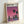 May include: Framed poster print leaning against white wall showing stylized cityscape silhouette with palm trees against gradient pink-purple sunset sky, water reflection below, in light wood frame on white surface