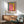 May include: Framed Rothko retrospective poster with colorful abstract painting featuring pink, yellow, and teal blocks mounted on white wall above mid-century modern wooden credenza. Scene shows gray upholstered armchair with wooden frame, round coffee table, and white ceramic table lamp creating cozy living room setting with natural sunlight casting shadows.