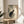 May include: Vintage Japanese woodblock print featuring a black cat sitting beneath flowering rose branches with green leaves, displayed in a thin gold frame on a round wooden side table next to a beige upholstered chair. Soft natural lighting creates shadows across the cream-colored wall, with a plant visible in the corner of the minimalist room setting.