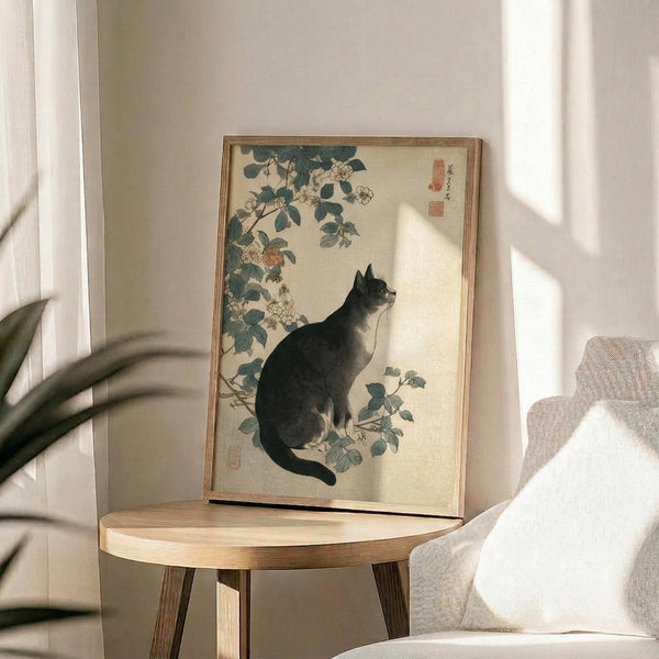 May include: Vintage Japanese woodblock print featuring a black cat sitting beneath flowering rose branches with green leaves, displayed in a thin gold frame on a round wooden side table next to a beige upholstered chair. Soft natural lighting creates shadows across the cream-colored wall, with a plant visible in the corner of the minimalist room setting.