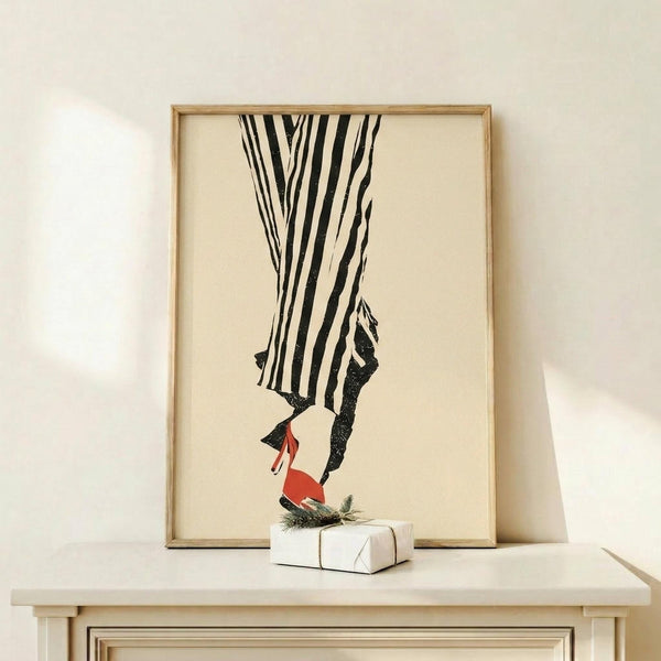 May include: Framed fashion illustration showing legs wearing black and white striped pants with red high heels, displayed on light wood console table next to small wrapped gift with greenery. Minimalist artwork in natural wood frame against cream wall in modern interior setting.