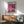 May include: Framed poster with pink-purple sunset sky, silhouetted palm trees, city skyline with lit buildings reflected in water, displayed above wooden sideboard with ceramic lamp in modern living room setting with gray upholstered chair and natural lighting creating window shadows on white wall.