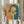 May include: Person holding framed vintage-style poster featuring woman with dark bob haircut smoking, rendered in teal and orange colors with Japanese text on cream background. Dried wheat stems visible in left hand. Soft natural lighting against neutral curtained backdrop.