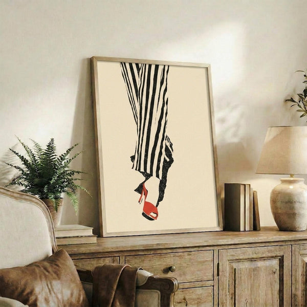 May include: Framed fashion illustration print displayed on rustic wooden sideboard in neutral-toned living room setting. Artwork shows minimalist line drawing of legs in black and white striped pants wearing red high heel shoes on cream background. Natural wood frame, surrounded by fern plant, ceramic vase, books, and beige upholstered furniture.