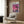 May include: Framed poster with purple-pink gradient sky, two dark palm trees, silhouetted city buildings, and water reflections, displayed above a beige upholstered armchair with geometric patterned pillow in modern living room with wood paneled walls, brass floor lamp, and side table with open book.