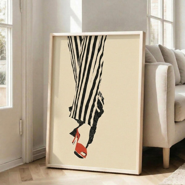 May include: Framed minimalist fashion artwork featuring black and white striped pants and red high heel shoes on cream background, displayed in light wood frame leaning against white wall next to beige upholstered sofa in bright living room with large windows