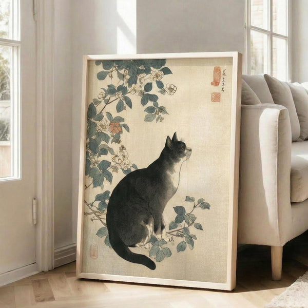 May include: Framed Japanese-style artwork featuring a black and white cat sitting among flowering rose branches with green leaves on cream background, displayed in light wood frame leaning against white wall next to beige upholstered sofa in bright living room with large windows