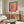 May include: Modern living room with cream walls featuring a framed colorful abstract art poster with pink, yellow, and blue blocks above a wooden sideboard. White curved chair and glass-top coffee table complete the mid-century modern setting with natural lighting.