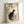 May include: A framed vintage Japanese woodblock print showing a black and white cat sitting among flowering rose branches with green leaves. The artwork features delicate botanical details on cream-colored paper with red artist seals. The print is displayed in a light wooden frame leaning against a white wall, creating soft shadows.