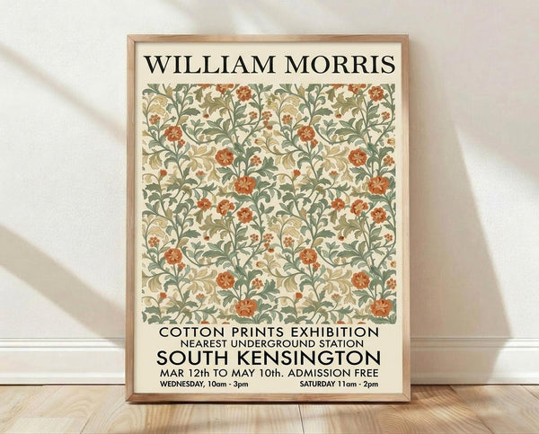 May include: Framed poster with ornate floral pattern in sage green, coral red, and cream colors featuring William Morris-style botanical design. Text reads "Cotton Prints Exhibition" and "South Kensington" with exhibition details. Light wood frame displayed against white wall on wooden floor with soft natural lighting creating shadows.