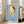 May include: Framed poster with light blue background featuring a stylized illustration of a young girl with brown hair, yellow headband, and yellow dress holding a small knife. The girl has large green eyes and pale skin in a minimalist art style. Text reads "YOSHITOMO NARA" with Japanese characters and dates "1992年8月31日〜10月31日". The wooden-framed artwork leans against white walls near a beige upholstered chair in a bright room with large windows.