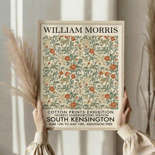 May include: Hands holding a framed poster featuring William Morris floral pattern in sage green and coral orange against cream background. Exhibition poster design with Cotton Prints Exhibition and South Kensington text. Light wood frame, neutral room setting with dried wheat stems and flowing curtains visible.