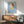 May include: Framed poster of stylized girl character with brown bob haircut, yellow halo and dress, holding small knife, on blue background with Yoshitomo Nara text below. Shown in modern living room setting above wooden sideboard with white ceramic lamp, next to mid-century green upholstered armchair with wooden frame. Natural sunlight creates dramatic shadows across white walls.